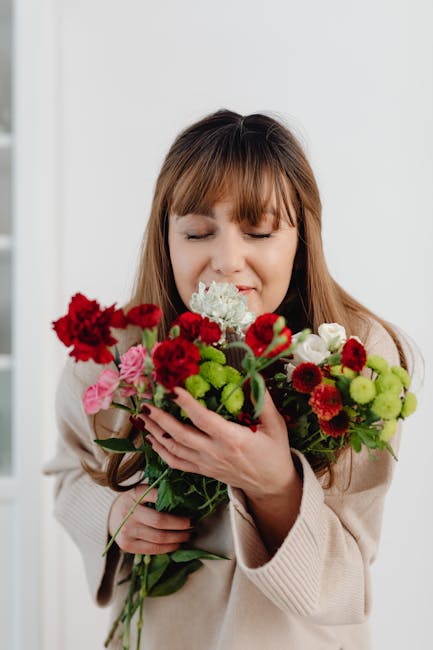 A woman with light brown hair and closed eyes holds a large bouquet of fresh flowers close to her face, appearing to smell the floral arrangement. The bouquet features a variety of flowers including r