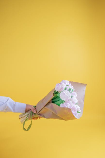 A person wearing a white shirt holds a bouquet of fresh, pastel pink tulips wrapped in light brown kraft paper with a green ribbon, against a bright yellow background. The tulips are fully bloomed wit