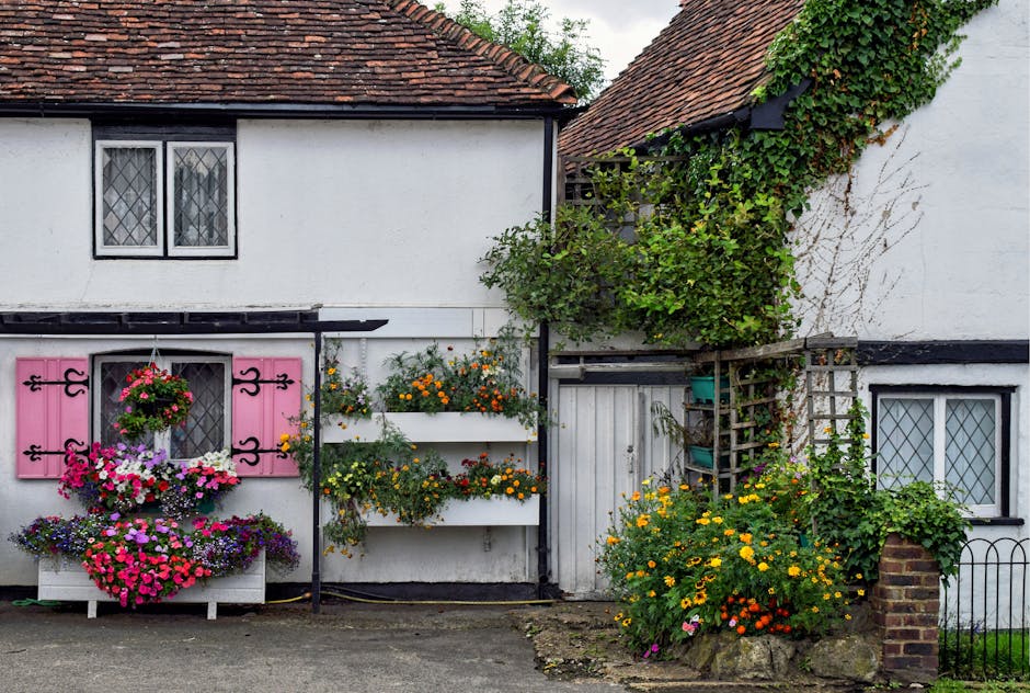 A quaint white cottage with black trim and a red tiled roof, adorned with vibrant hanging baskets and window boxes filled with colorful flowers such as pink petunias, purple pansies, orange marigolds,